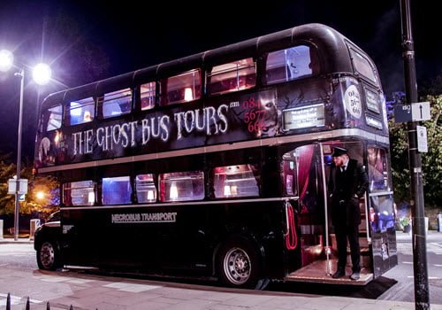 red bus at night lit up with conductor standing on edge of bus at back